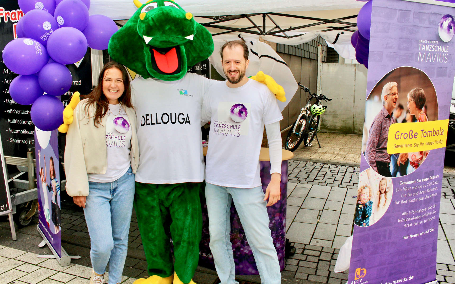 Maskottchen Dellouga mit zwei Personen an einem Infostand am Kirmessonntag