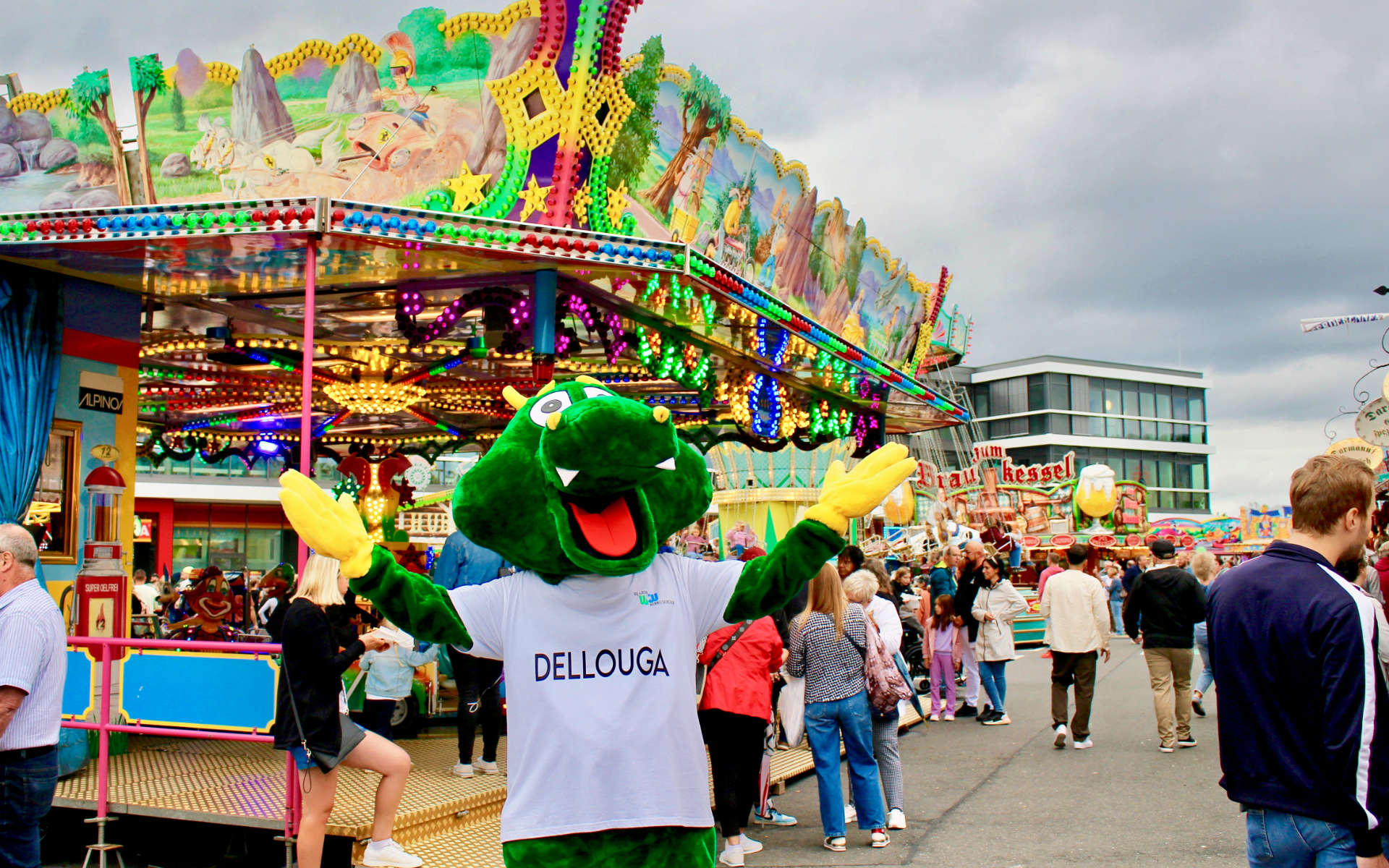 Maskottchen Dellouga vor einem Karussell auf der Kirmes